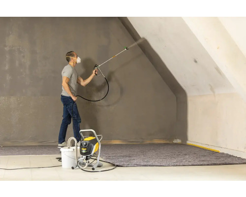 En man sprutar färg med en färgspruta på en vägg i ett rum.