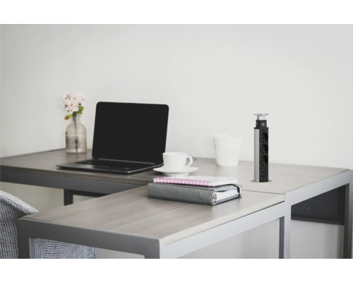 Arbetsrum med bärbar dator, kaffekopp, anteckningsböcker och infällbar uttagslist på ett skrivbord.