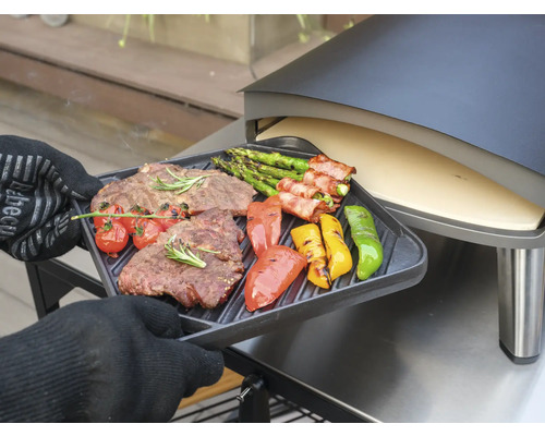 En person med ugnsvantar håller en grillbricka med grillad biff, sparris, tomater och paprika framför en pizzaugn.