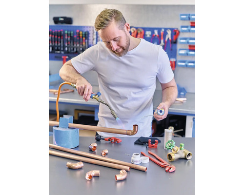 En man löder ett kopparrör med en lödkolv. Kopparrör, beslag och verktyg ligger på bordet.