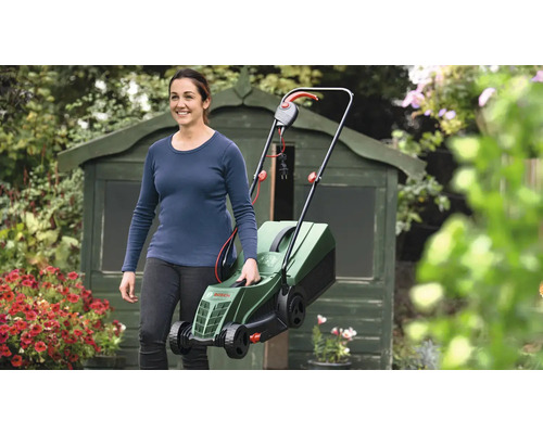 Kvinna bär en elektrisk gräsklippare i trädgården framför ett redskapsskjul.