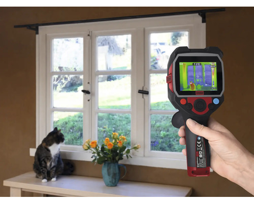 En hand håller en värmekamera framför ett fönster, medan en katt sitter på fönsterbrädan.