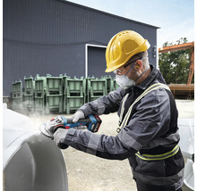 En man med skyddshjälm, skyddsglasögon och andningsskydd slipar ett arbetsstycke med en batteridriven vinkelslip.