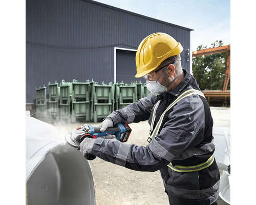 En man med skyddshjälm, skyddsglasögon och andningsskydd slipar ett arbetsstycke med en batteridriven vinkelslip.