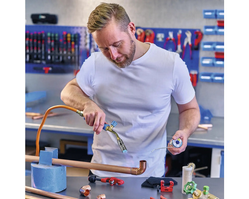 En man löder kopparrör med en lödkolv i en verkstad, med kopparfittings och verktyg på bordet.