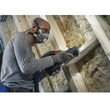 Man arbetar med multifunktionsverktyg på träkonstruktion med dammutsug, andningsskydd, skyddsglasögon och öronproppar för arbetarskydd