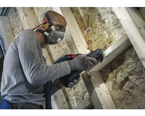 Man arbetar med multifunktionsverktyg på träkonstruktion med dammutsug, andningsskydd, skyddsglasögon och öronproppar för arbetarskydd