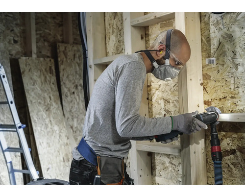 Hantverkare med andningsskydd och skyddsglasögon som arbetar med ett multifunktionsverktyg i ett skalbyggnad