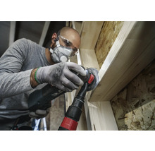 Hantverkare bearbetar trä med ett multifunktionsverktyg, inklusive dammutsugning, skyddsglasögon och andningsskydd.