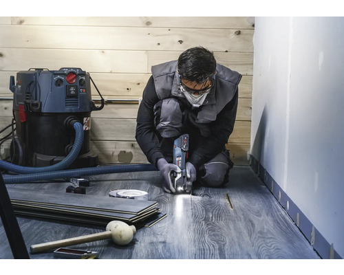 En man knäböjer och skär laminat med ett multifunktionsverktyg. En dammsugare, laminatgolv och verktyg är också synliga.