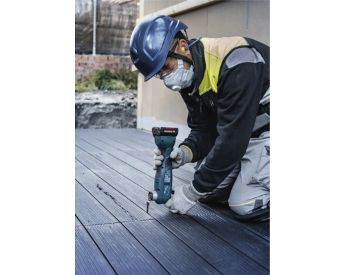 En byggnadsarbetare med skyddshjälm och ansiktsmask använder ett multifunktionsverktyg för att skära ett spår i en trädgårdsplatta.