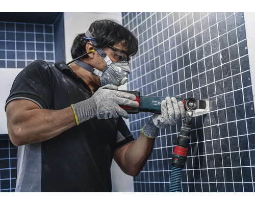 Man tar bort kakel med ett multiverktyg, utrustad med skyddsglasögon, hörselskydd, dammmask och handskar