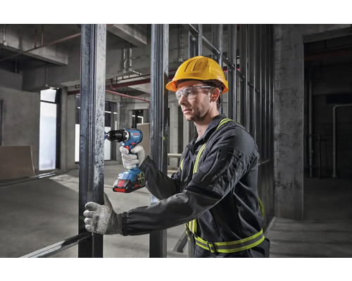 En arbetare med skyddshjälm och skyddsglasögon använder en batteridriven skruvdragare på en byggarbetsplats.