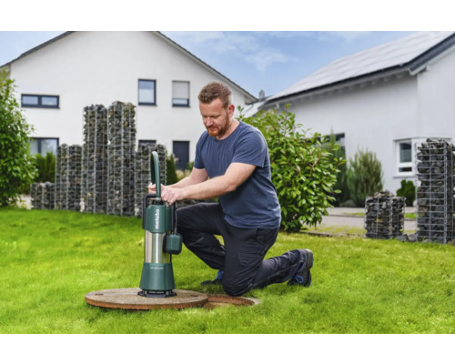 En man använder en Metabo dränkbar pump på en gräsmatta framför ett hus. Pumpen används för vattenavlägsning.