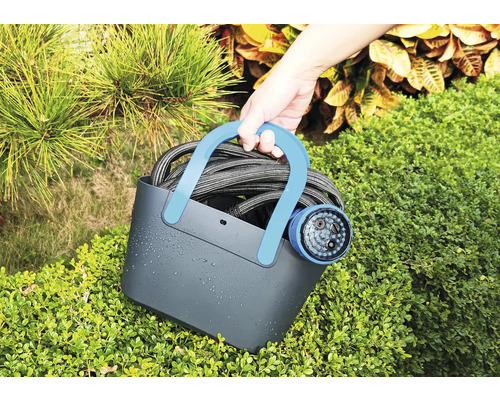 Person håller en vattenslang med trädgårdsspruta i en bärkorg framför buskar.