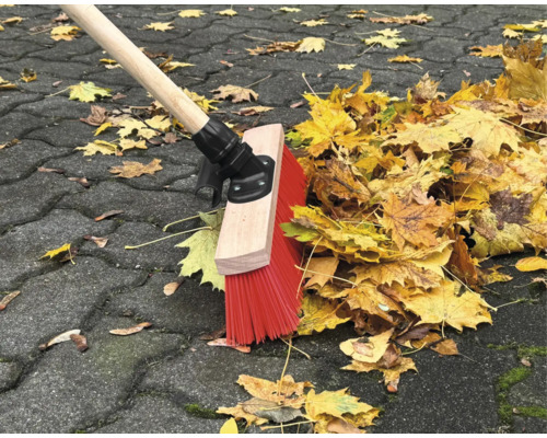 En sopmaskin med trähandtag och röda borst sopar löv på en stenlagd gångväg.