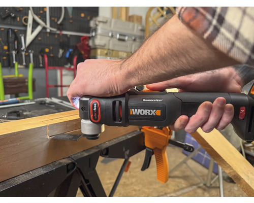 En person använder ett Worx multiverktyg för att bearbeta en träbit. Verktyget är svart och orange.