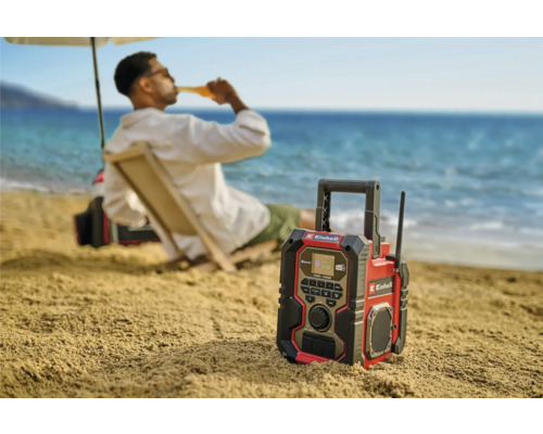 En Einhell byggnadsradio på stranden med en person i bakgrunden som njuter av en drink.