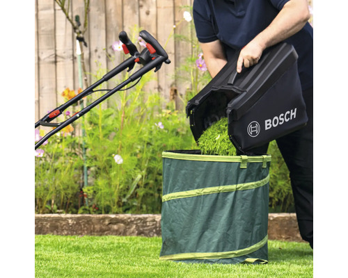 En person tömmer gräsklipp från en gräsklippares uppsamlare i en trädgårdssäck. En gräsklippare syns i bakgrunden. Bosch logotyp.