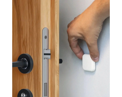 En hand fäster ett magnetiskt dörrstopp på en vägg bredvid en trädörr med ett silverfärgat dörrlås.