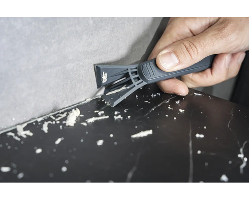 En person tar bort silikonfogar mellan vägg och yta med en fogskrapa.