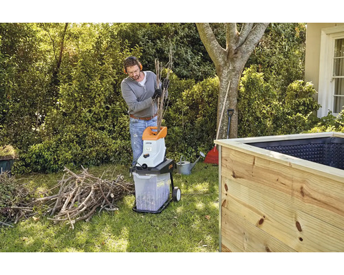 En man använder en trädgårdshackare för att strimla grenar bredvid en upphöjd rabatt.