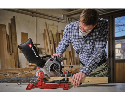 En man med skyddsglasögon och andningsskydd sågar en träplanka med en Einhell kap- och geringssåg.