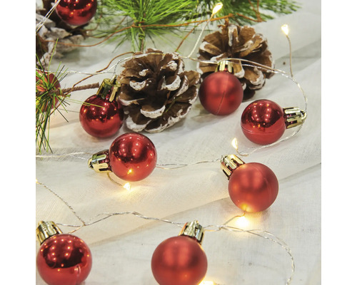 Dekorativt arrangemang med röda julgranskulor, ljusslinga, tallkottar och grangrenar.