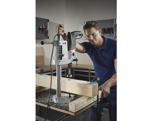 Man borrar ett hål i en träbit med en bänkborrmaskin. Han bär skyddsglasögon.