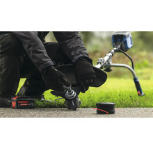 En person byter blad på en grästrimmer med ett batteri och en spole i närheten.