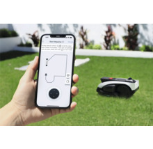 Person håller smartphone med app för att styra en gräsklipparrobot. I bakgrunden finns gräsklipparroboten på en gräsmatta.