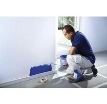 En man målar en vägg med en färgroller i ett rum. Maskeringstejp skyddar golvsockeln och golvet.