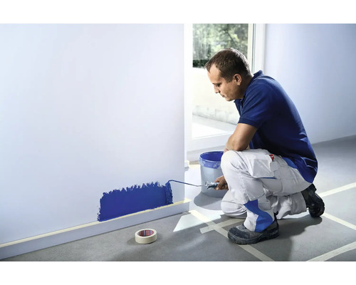 En man målar en vägg med en färgroller i ett rum. Maskeringstejp skyddar golvsockeln och golvet.