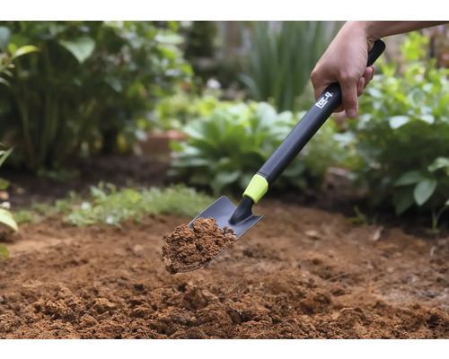 En handspade med jord hålls mot en trädgårdsbakgrund