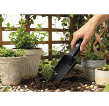 Trädgårdsscen med blomkrukor, omgivande grönska och en person som använder en planteringsspade.