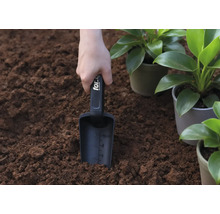 En hand håller en planteringsspade i jorden, bredvid krukväxter.