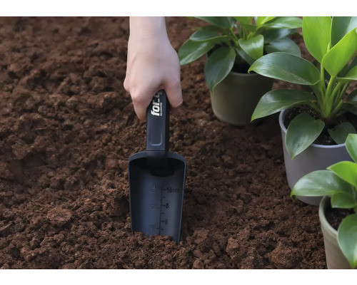 En hand håller en planteringsspade i jorden, bredvid krukväxter.