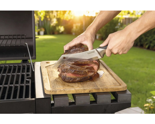 Tillagad biff skärs med en kniv på en träbräda bredvid en grill.