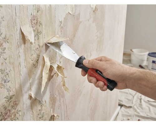 En person tar bort gammal tapet från en vägg med en spackelspade.
