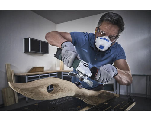 Användare med andningsskydd, skyddsglasögon och handskar bearbetar trä med en vinkelslip