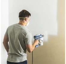 Man målar vägg med färgspruta, utrustad med skyddsglasögon och dammmask