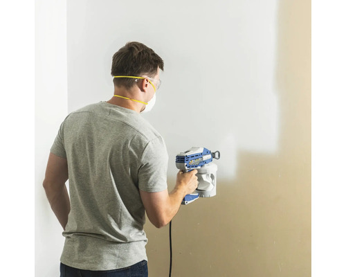 Man målar vägg med färgspruta, utrustad med skyddsglasögon och dammmask