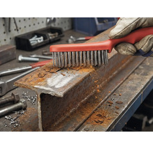 En hand med arbetshandske rengör en rostig stålprofil med en stålborste.