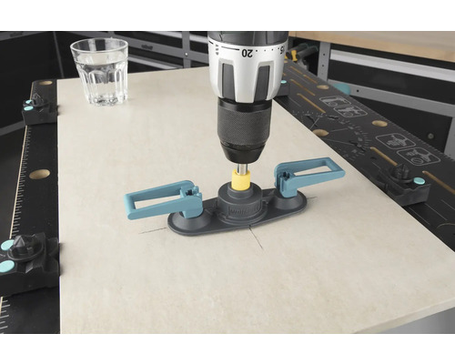 En borrmaskin med kakelborrhjälp borrar i en kakelplatta, ett glas vatten står bredvid.