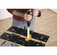 En person borrar med en borrmaskin i en träbit som är fäst på en arbetsbänk med ett gult fästelement.