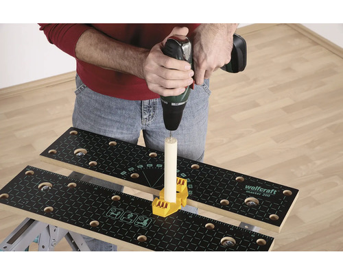 En person borrar med en borrmaskin i en träbit som är fäst på en arbetsbänk med ett gult fästelement.