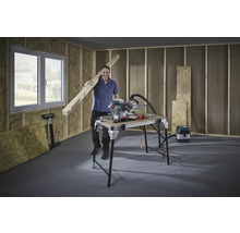 Man med träbjälke, kap- och gersåg och arbetsbänk i ett oinrett rum.