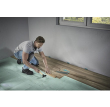 En man lägger laminatgolv med en hammare och slagkloss på stegljudsisolering i ett rum.