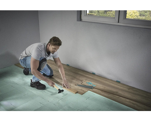 En man lägger laminatgolv med en hammare och slagkloss på stegljudsisolering i ett rum.