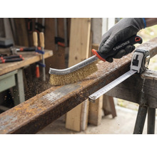 En person använder en stålborste för att ta bort rost från en metallbit bredvid ett rullmått.